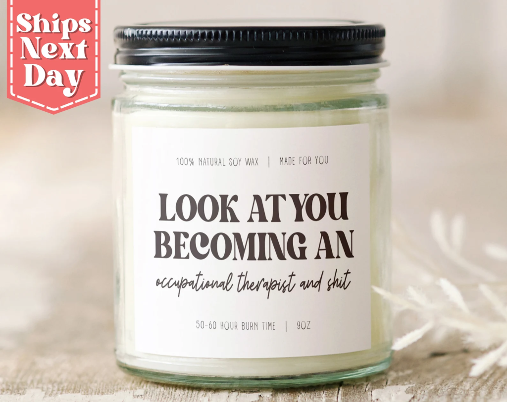 Humorous soy wax candle in a glass jar with a black lid. The label reads, “LOOK AT YOU BECOMING AN occupational therapist and shit,” with smaller text stating “100% natural soy wax | Made for you” and “50–60 hour burn time | 9 oz.” A red badge in the top left corner says “Ships Next Day.”
