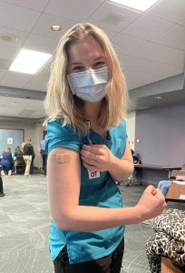 Clarice in her hospital scrub and a mask flexing her arm and showing off bandaid after receiving the COVID-19 vaccine
